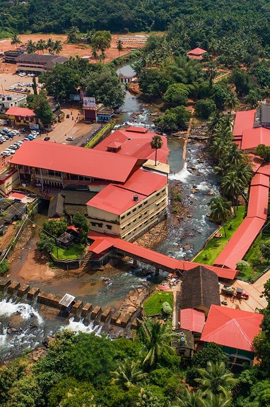 Aerial view of red-roofed buildings beside a rocky river surrounded by coconut palms near Mangaluru